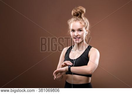 Happy young blond sportswoman in activewear and earphones looking at you while switching on her fitbit before outdoor workout