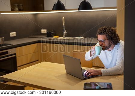 Male Freelancer Sitting At Dinning Room Table, Wearing Pajamas, Drinking Coffee And Working Late Usi