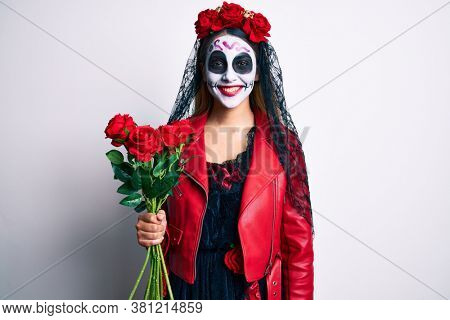 Woman wearing day of the dead costume holding roses looking positive and happy standing and smiling with a confident smile showing teeth 