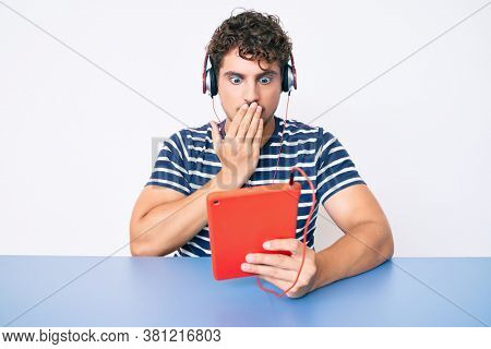 Young caucasian man with curly hair using touchpad sitting on the table covering mouth with hand, shocked and afraid for mistake. surprised expression 