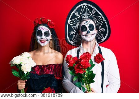 Couple wearing day of the dead costume holding flowers looking positive and happy standing and smiling with a confident smile showing teeth 