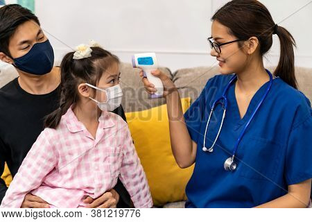 Asian female doctor use infrared thermometer to take temperature to little girl child sit with her dad in living room while doctor visit at home. Home health care delivery and doctor visiting concept.