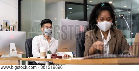 Panorama asian Office employee with protective face mask working in new normal office with social distance practice to american african colleague in foreground prevent coronavirus COVID-19 spreading.