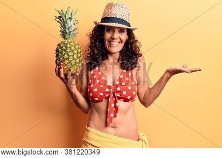 Middle age beautiful woman wearing bikini and summer hat holding pineappel celebrating achievement with happy smile and winner expression with raised hand 