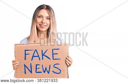 Young beautiful blonde woman holding fake news banner looking positive and happy standing and smiling with a confident smile showing teeth 
