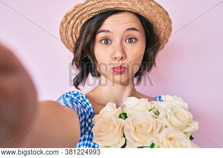 Young beautiful girl wearing summer hat taking a selfie puffing cheeks with funny face. mouth inflated with air, catching air. 