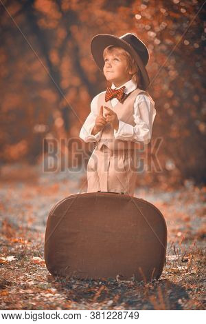 Happy little boyboy in a classic suit and hat possing with a large brown retro suitcase in a beautiful autumn park. Retro style. Children's fashion.