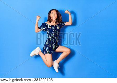 Young beautiful chinese woman wearing casual clothes smiling happy. Jumping with smile on face doing strong geture over isolated blue background
