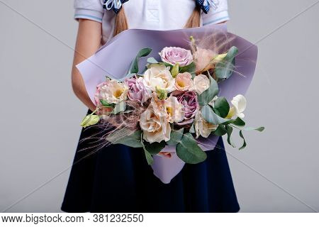 A Bouquet Of Flowers, Packed In Purple Paper In The Hands Of A Girl With Pigtails On A Light Backgro