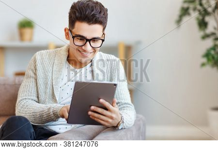 Delighted Young Ethnic Male In Glasses Smiling And Browsing Social Media On Tablet While Chilling On