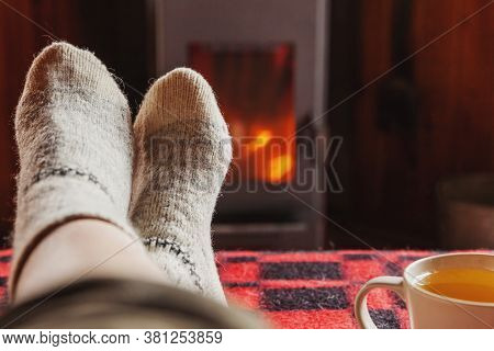 Feet Legs In Winter Clothes Wool Socks And Cup Tea At Fireplace Background. Woman Sitting At Home On
