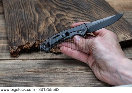 Small Folding Knife In Hand. The Blade And Knife Handle Are Black. On A Wooden Background.