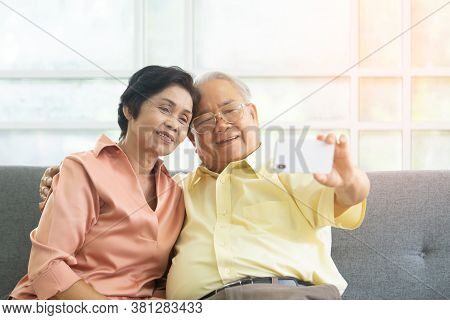 Happy Senior Asian Couple Is Maiking Video Call On Mobile Phone On Sofa At Home.