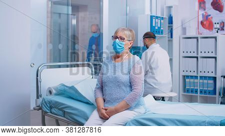 Portrait Of Elderly Lady Sitting On The Hospital Bed During Covid-19 Crisis. Doctor And Patients Wea