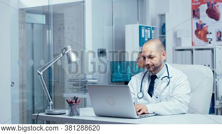 Doctor Typing An E-mail From Modern Office Of Private Clinic With Glass Walls, Nurse In Background C