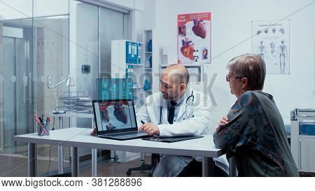 Young Male Medic Presenting A Heart Booklet On Pc To Elderly Retired Senior Woman Patient. Heart Dis