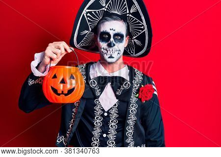 Young man wearing mexican day of the dead costume holding pumpkin looking positive and happy standing and smiling with a confident smile showing teeth 