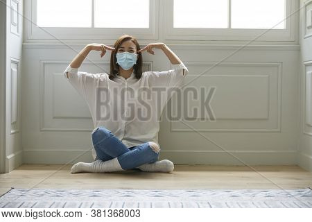 Young Asian Woman In Quarantine At Home Wearing Facial Mask Sitting On Floor Legs Crossed Looking Ha