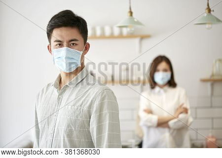 Portrait Of A Young Asian Man Wearing Mask Looking At Camera With Husband In The Background