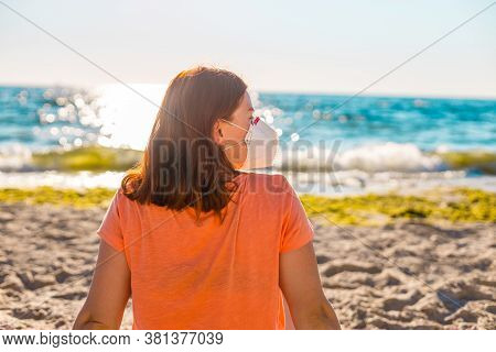 Young Tourist Girl In A Medical Mask With Her Back Sits On The Beach Near The Sea. New Rules To Wear