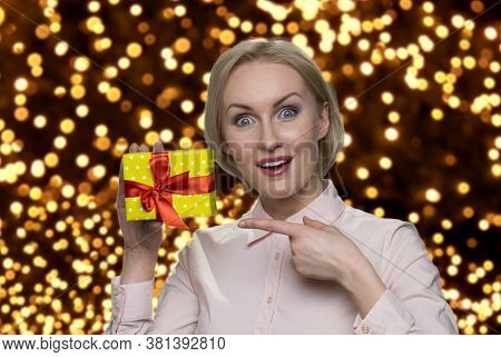 Happy Shocked Woman Pointing At Gift Box. Excited Woman Showing Peresent Box. Golden Bokeh Lights On