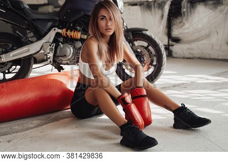 Attractive Sportswoman Posing In A Light Studio Sitting On The Floor With A Large Orange Punching Ba