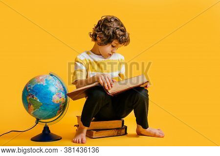 Curly Boy In Glasses And Stripes Tshirt And Sits On A Stack Of Books, Near To The Globus On Yellow B