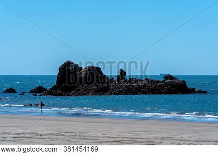 A Huge Rock Outcrop Jutting Out Of The Sea Near The Beach Where A Lone Fisherman Stands