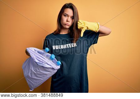 Beautiful volunteer girl caring environment doing volunteering holding bag with rubish bottles with angry face, negative sign showing dislike with thumbs down, rejection concept