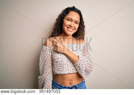 Young beautiful woman with curly hair wearing casual t-shirt standing over white background cheerful with a smile of face pointing with hand and finger up to the side with happy and natural expressio
