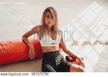 Attractive Sportswoman Posing In A Light Studio Sitting On The Floor With A Large Orange Punching Ba