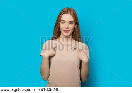Confident Caucasian Woman With Red Hair And Freckles Posing On A Blue Wall Is Pointing To Her And Lo