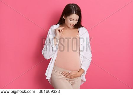 Pregnant Woman Standing Isolated Over Rose Background Touching Her Belly And Looking At It, Wearing 