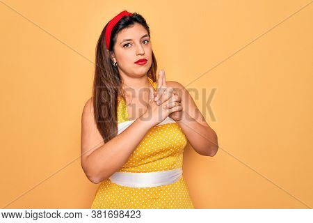 Young hispanic pin up woman wearing fashion sexy 50s style over yellow background Holding symbolic gun with hand gesture, playing killing shooting weapons, angry face
