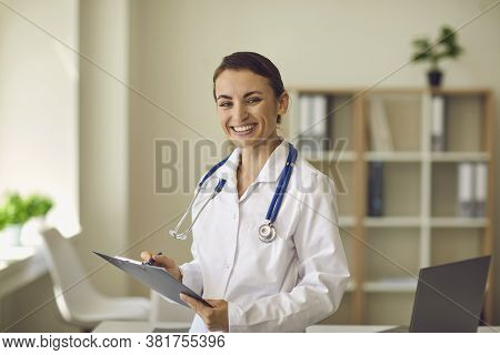Young Smiling Doctor Therapist Looking At Camera And Making Notes Over Medical Clinic Office At Back