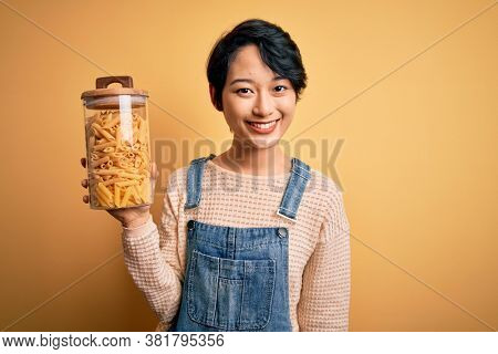 Young beautiful asian girl holding jar with italian dry pasta macaroni over yellow background with a happy face standing and smiling with a confident smile showing teeth