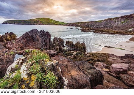 Small Sandy Hidden Beach Between Cliffs With View On Dingle Bay And Lighthouse. Dramatic Sunset In D