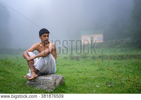 Young Man Wearing A Dhoti, Sitting On A Rock, Open In The Green Field In A Foggy Morning.