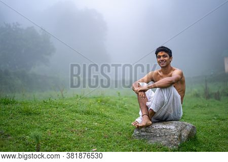Young Man Wearing A Dhoti, Sitting On A Rock, Open In The Green Field In A Foggy Morning.