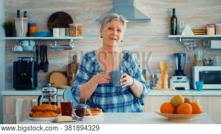 Happy Senior Woman Holding Cup Of Coffee While Looking At Camera In Ktichen During Breakfast. Authen