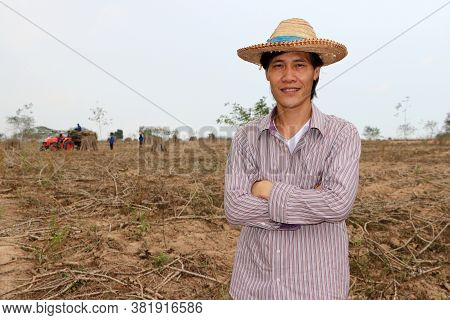 Male Farmer Standing And Hugging Chest In The Cassava Farm With Out Focus Tractor And Worker.
