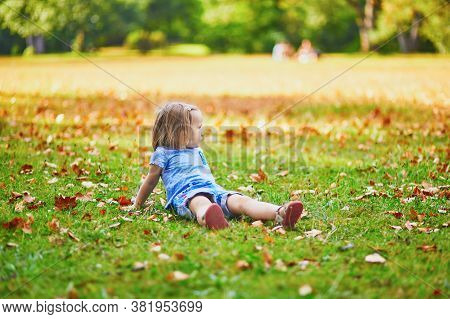 Unhappy And Emotional Toddler Girl Sitting On The Ground In Park. Misbehaving Child On The Street. T