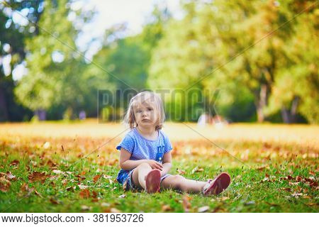 Unhappy And Emotional Toddler Girl Sitting On The Ground In Park. Misbehaving Child On The Street. T