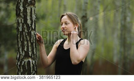 Young Caucasian Woman In The Nature. Listening The Music On The Wireless Headphones And Touching The
