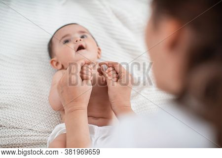 Cute Baby Sitting With Support On Bed. Mother Supports Child Sitting, Spreading Her Hands To Side. C