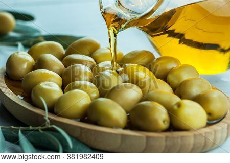 Pouring Homemade Olive Oil From Bottle Into Glass On Raw Turkish Green Olives In Bamboo Bowl, Olivae