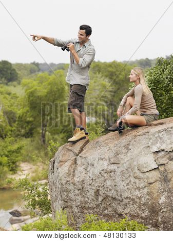 Vista lateral de comprimento total de um homem jovem, apontando para o vista por mulher em pedra com binóculos