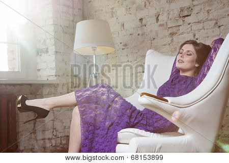 Fashion glamour girl dreamily sitting in white chair in violet lace dress.