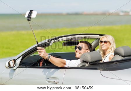 road trip, leisure, couple, technology and people concept - happy man and woman driving in cabriolet car and taking picture with smartphone on selfie stick