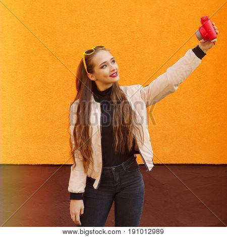Young pretty hipster girl in yellow sunglasses make selfie by pink instant camera on orange wall.